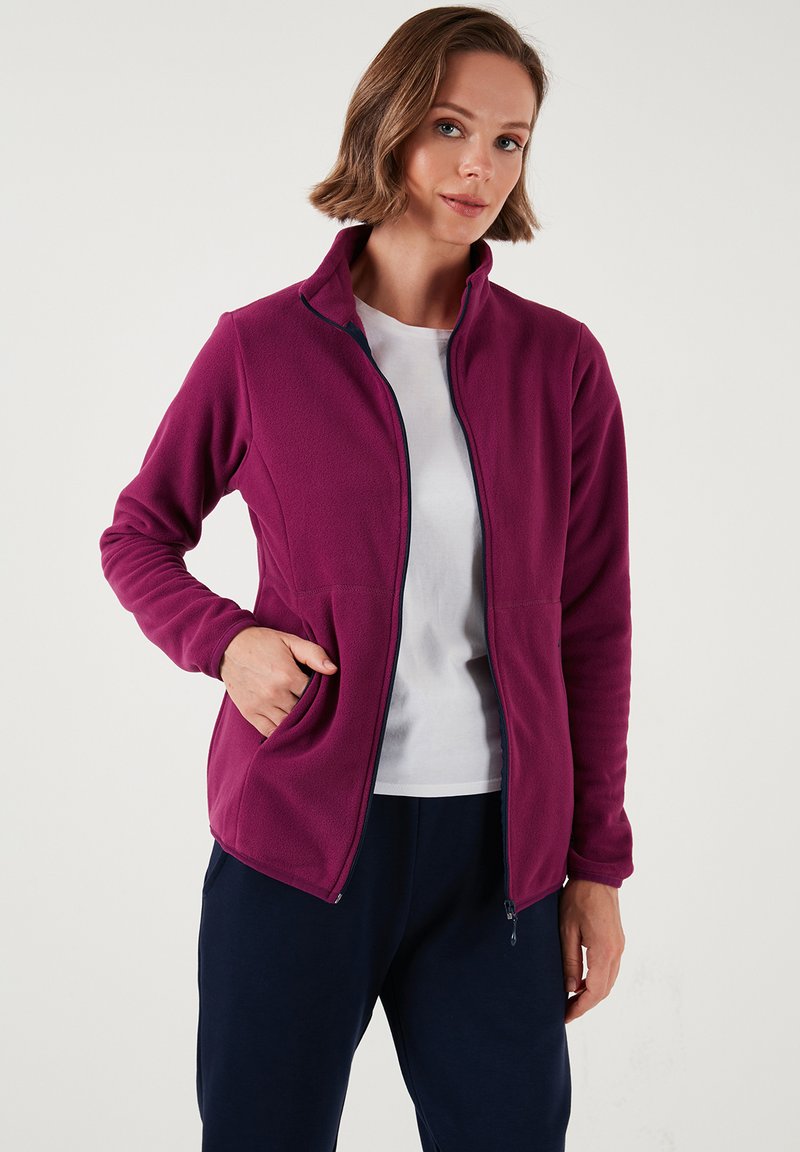 Flis jakna tamno ružičaste boje s visokim ovratnikom, zatvaranjem na patentni zatvarač i bočnim džepovima. Glatka textura i slim fit dizajn. Nosena preko bijele košulje.