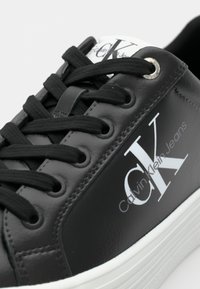 Zapatilla sintética negra con logo blanco, diseño con cordones, textura suave y suela blanca contrastante. Presenta ojales de metal y logo en la lengüeta.
