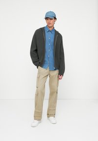 Felpa grigia a costine con zip, camicia di jeans azzurra con bottoni, pantaloni cargo beige con tasche e sneakers bianche. Un cappellino blu completa l'outfit.