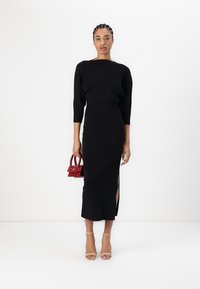 Robe noire en tricot avec des manches trois-quarts, fente sur le côté, portée avec des talons nus et un petit sac à main rouge. Texture lisse et design ajusté.