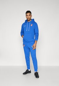 Blauer Hoodie und passende Jogginghosen aus weichem Stoff, mit einem Logo und weißen Akzenten. Schwarze Sportschuhe runden das Outfit ab.