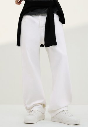 Személy, aki laza fehér nadrágot, fehér tornacipőt visel, és fekete pulóvert kötött a derekára, egyszínű háttér előtt áll.