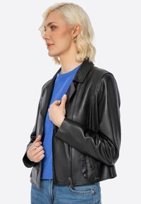 Schwarze Lederjacke mit taillierter Passform, Reißverschluss und Seitentaschen. Über einem blauen Pullover getragen, der eine elegante Textur und einen glänzenden Finish zeigt.