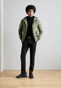 Veste matelassée verte avec capuche, portée sur un col roulé noir, accompagnée de pantalons noirs fuselés et de chaussures noires à lacets. Design simple et fonctionnel.