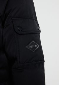 Svart boblejakke med ermer som har en lomme med en svart trykknapp og et diamantformet svart logo-lapp preget med "REPLAY".