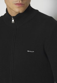 Schwarzer Reißverschluss-Pullover mit strukturiertem Strickdesign und hohem Kragen, mit einem weißen "GANT"-Logo auf der linken Seite.