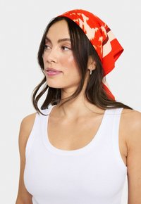 Rød mønstret tørklæde lavet af glat stof, med abstrakte designs og en knude bagpå, båret med en hvid tanktop.