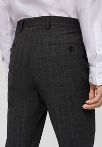 Pantalons à carreaux gris en tissu tissé, avec un devant plat, des poches latérales et une poche arrière à bouton. Détail de conception texturée visible.