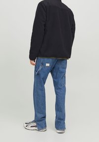 Zwarte fleecejas met gestructureerde stof, gecombineerd met lichtblauwe denim cargobroek met zakken en een labeldetail. Witte sneakers.