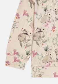 Tissu couleur crème avec un motif floral comprenant des lapins, des fleurs roses et des feuilles vertes. Texture douce, design de manches détendues.