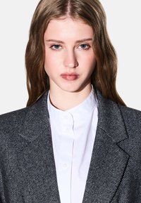 Blazer grigio a righe con rever a punta, indossato sopra una camicia bianca con bottoni. Tessuto liscio, design sartoriale con spalle strutturate.