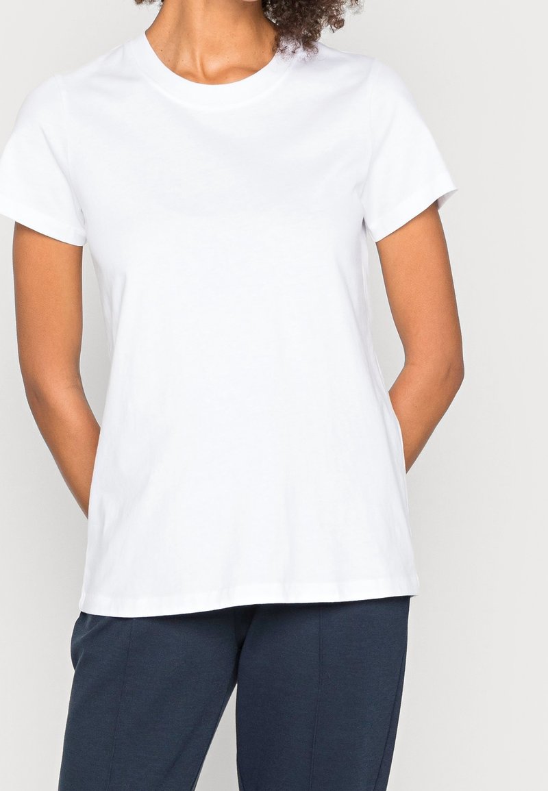 Personne portant un T-shirt blanc uni à manches courtes et un pantalon bleu foncé, debout les mains derrière le dos.