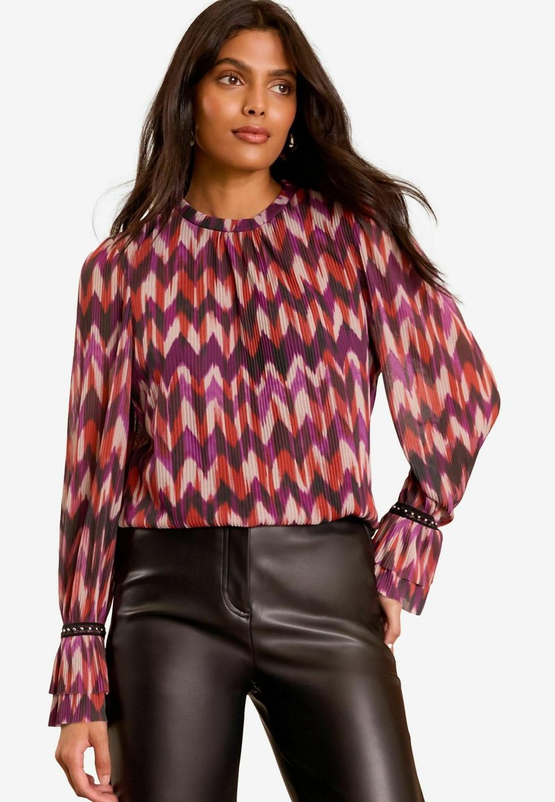 Mehrfarbige Langarmbluse mit plissiertem Design, das ein Zickzackmuster in Rosa, Rot und Lila zeigt. Schwarze Hosen mit einem eleganten Finish.