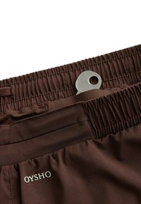 Short marron à taille élastique avec une petite clé argentée partiellement glissée dans la poche près de l’étiquette de la marque Oysho.