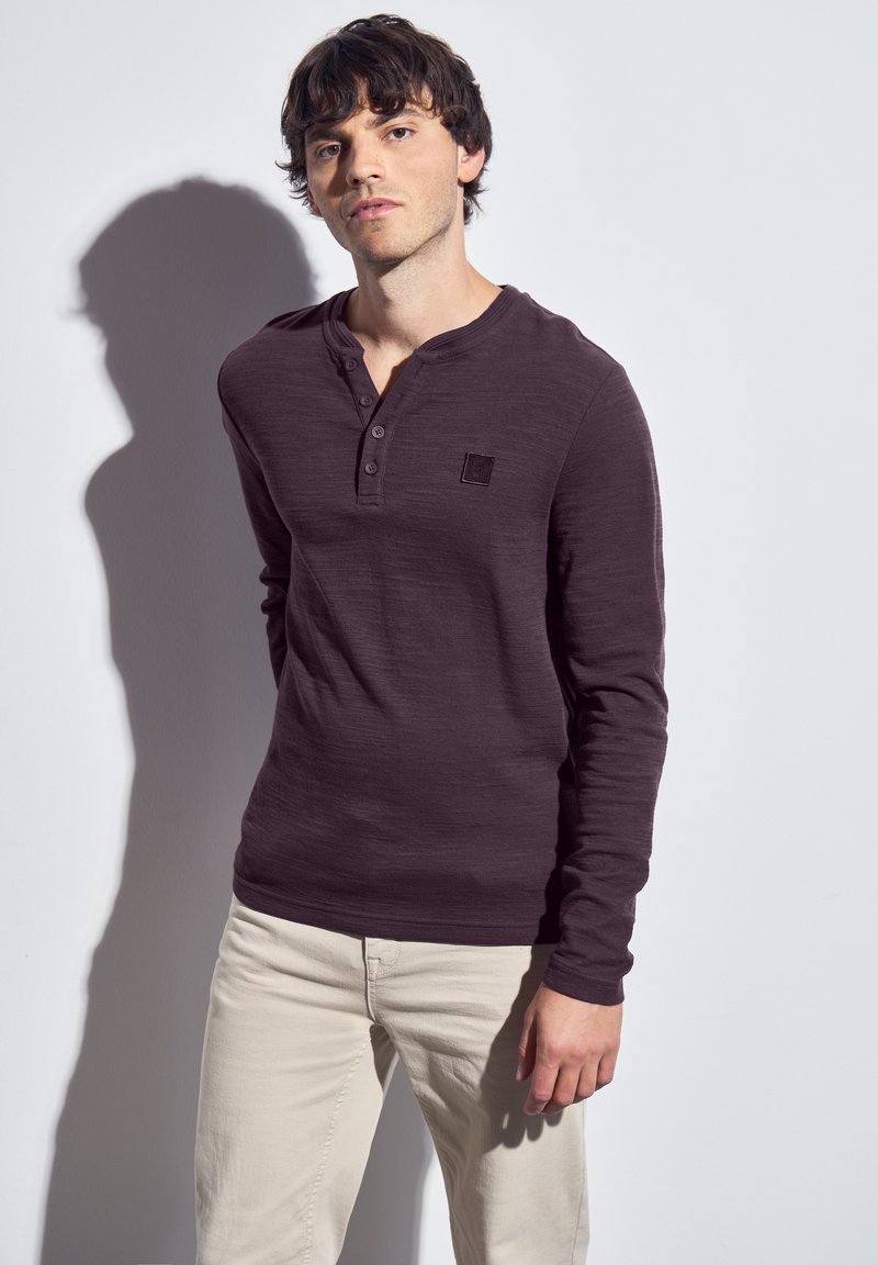 Long-sleeve, dark purple henley shirt with three buttons, textured fabric, and a small black logo patch on the chest. Worn with light beige pants.