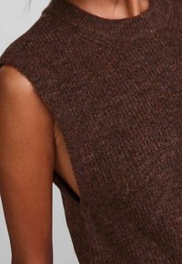 Brun ribbad stickad ärmlös topp med hög halsringning och öppna sidor, som visar upp ett strukturerat tyg.