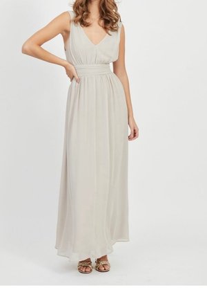 Woman wearing sleeveless, light beige, floor-length dress with V-neck and wide waistband, paired with gold braided sandals.