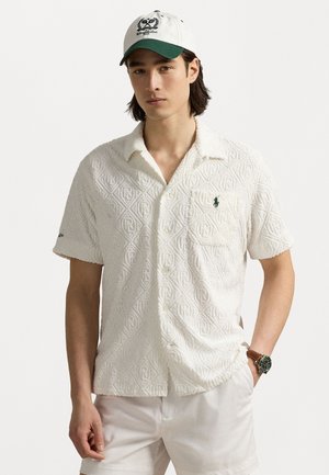 Witte korte mouwen shirt met een gestructureerd ruitpatroon. Heeft een groen logo op de borstzak en een kraag. Pet op de achtergrond.