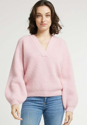 Pull en tricot rose clair avec un col en V, des manches surdimensionnées et une texture côtelée. Associé à un jean bleu, mettant en valeur une silhouette décontractée.