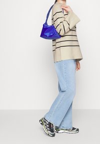Beige gestreepte oversized trui, lichtblauwe jeans en zwarte sportschoenen met groene accenten. Houdt een kleine glanzende blauwe handtas vast.