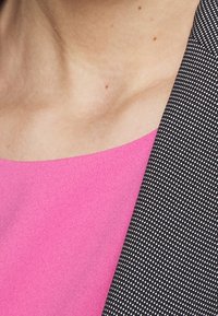 Haut fuchsia à texture lisse, associé à un blazer noir avec un petit motif à pois, mettant en valeur les contrastes de couleurs et de designs.