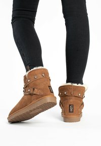 Braune Wildleder-Stiefeletten mit einem Kunstfellfutter, Nietenapplikationen und einem verstellbaren Riemen. Strukturierte Gummisohle mit einem Rautenmuster.