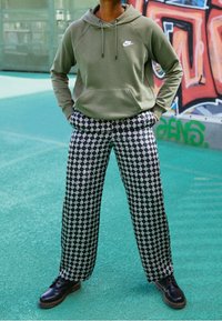 Oliwkowa zielona bluza z kapturem z czarnym logo, zestawiona z szerokimi spodniami w czarno-biały wzór. Całość dopełniają czarne buty na sznurowadła.