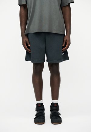 Man draagt donkergrijze korte broek, grijs oversized T-shirt, witte sokken en zwarte hoge sneakers met rood logo, staand tegen een witte achtergrond.