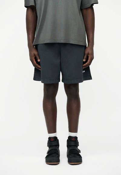 Homme portant un short gris foncé, un T-shirt gris oversize, des chaussettes blanches et des baskets montantes noires avec un logo rouge, debout devant un fond blanc.