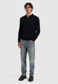 Zwarte zip-nektrui met lange mouwen, gecombineerd met versleten lichtblauwe jeans. Zwarte schoenen maken de outfit compleet. Effen achtergrond.