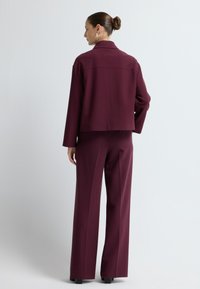 Bordeaux op maat gemaakt pak met een getailleerde blazer en wijde broek. Glad stof, minimalistisch ontwerp en een detail aan de achterkraag.