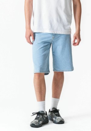 Muž oblečený v svetlomodrých rifľových kraťasoch, bielych ponožkách, čierno-šedých bežeckých topánkach a bielom tričku na bielom pozadí.