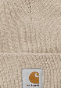 Berretto lavorato a maglia beige con texture a costine. Presenta un'etichetta quadrata bianca con un logo arancione stilizzato e il nome del marchio in basso.