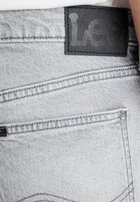 Γκρι τζιν παντελόνι με μαύρη δερμάτινη ετικέτα στη μέση που φέρει ανάγλυφο κείμενο "Leo". Περιλαμβάνει ορατές ραφές και μια πίσω τσέπη.