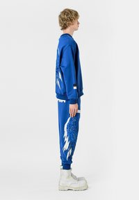 Sudadera azul y pantalones de chándal a juego que presentan grandes diseños de alas en blanco y azul. Hechos de una tela suave, con botas blancas de caña alta.