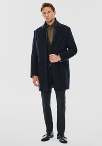 Granatowy wełniany płaszcz z dużymi klapami, zapięciem na guziki i bocznymi kieszeniami, nałożony na ciemnozieloną koszulę i dopasowane czarne spodnie.