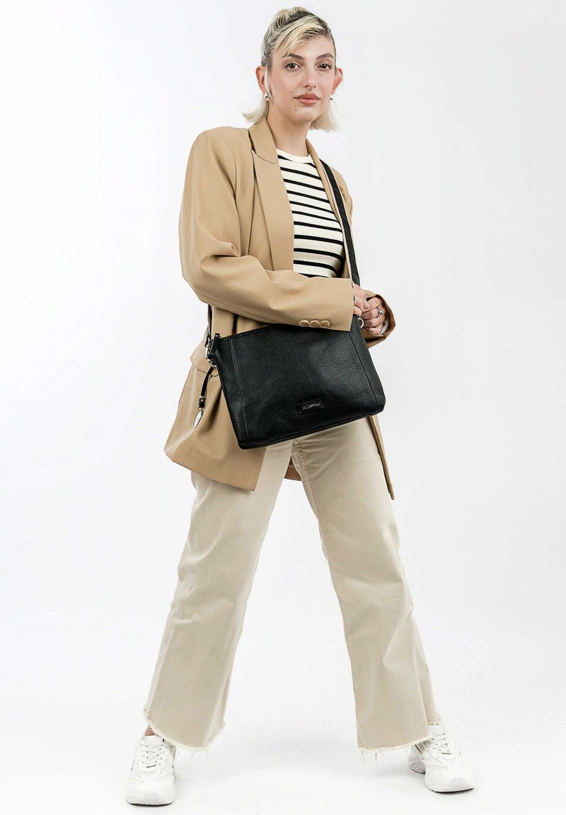 Schwarze Leder-Umhängetasche mit strukturierter Oberfl�äche, rechteckiger Form und silbernem Beschlag, getragen von einer Person in einem beigen Blazer und einem gestreiften Hemd.