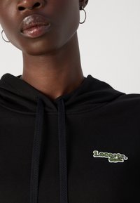 Un sweat à capuche noir avec un cordon de serrage, comportant un logo Lacoste brodé vert sur la partie gauche de la poitrine. Tissu doux et détails côtelés.