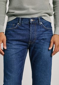Jeans de mezclilla azul oscuro con corte recto, cinco bolsillos y costuras amarillas en contraste. Cuenta con un cierre de botón de metal y cremallera.