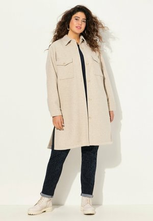 Beige wool blend coat with a button closure, two front pockets, and a relaxed fit, paired with navy patterned pants and cream ankle boots.
