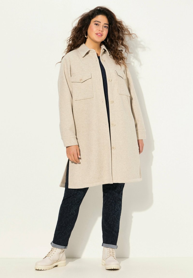 Beige kabát z vlněné směsi s knoflíkovým zapínáním, dvěma předními kapsami a volným střihem, spojený s námořnickými vzorovanými kalhotami a krémovými kotníkovými botami.