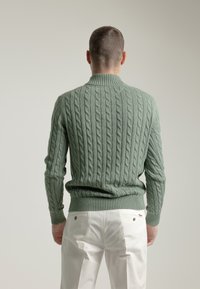 Suéter verde de punto trenzado con cuello alto, dobladillo y puños acanalados, combinado con pantalones blancos, mostrando una vista trasera texturizada.
