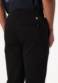 Juodos sportinės kelnės su minkšta tekstūra, elastinga liemene ir užtrauktuku užsegama užpakaline kišene. Ant liemenės esantis mažas geltonas logotipo ženklelis.
