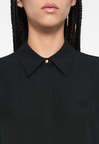 Fekete galléros ing sima anyagból, gombos lezárással. Bal mellrészen dombornyomott logóval és elegáns kialakítással.