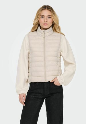 Beige puffer vest with horizontal quilted pattern, high collar, and front zipper, paired with cream balloon-sleeved top and black wide-leg pants.