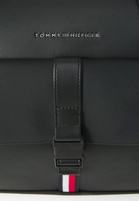 Black leather messenger bag with a textured surface, featuring a buckle closure and a prominent brand logo. Accented with red, white, and blue stripes.