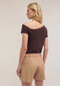 Top scurt de culoare maro, cu umerii descoperiți, asociat cu pantaloni scurți din suede bej deschis. Materialele sunt moi, cu un design ajustat și detalii minime.