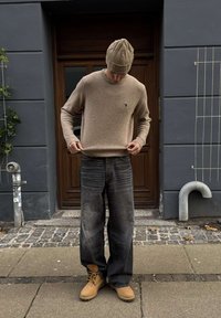 Muž s béžovou pletenou čiapkou, béžovým svetrom, voľnými tmavými džínsami a hnedými čižmami stojí na chodníku z kockového kameňa pred drevenými dverami.