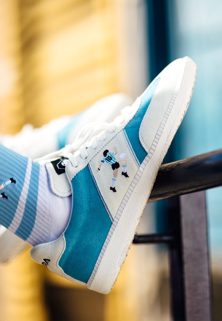 Blauwe en witte sneakers met een bovenwerk van suède, voorzien van een geborduurde voetballerontwerp aan de zijkant, aangevuld met gestreepte sokken.