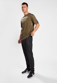 Olivgrünes Sport-T-Shirt mit kurzen Ärmeln, kombiniert mit schwarzen, schmal zulaufenden Hosen. Das Outfit zeichnet sich durch eine glatte Textur und ein leichtes Design aus.